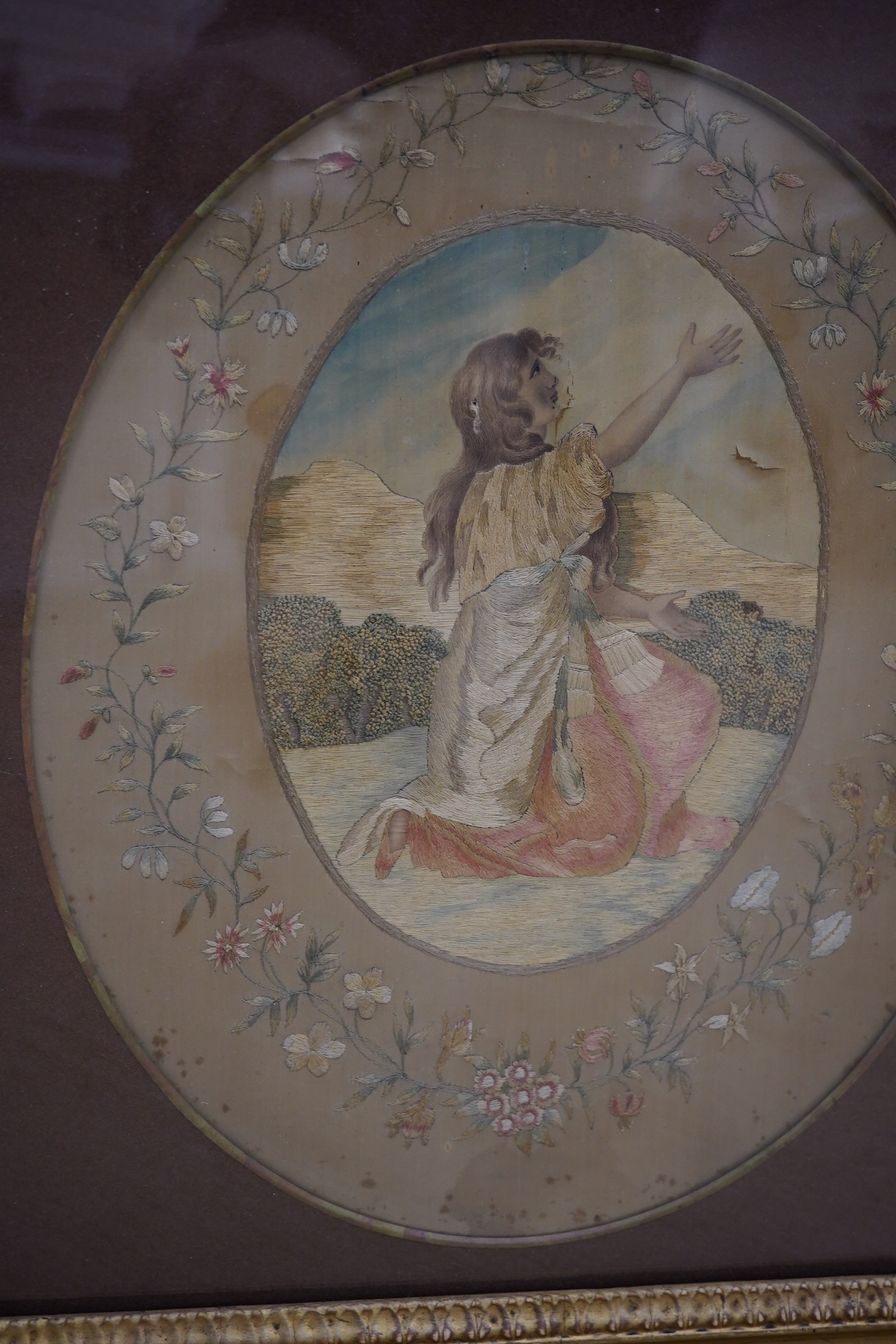 A Regency oval silkwork embroidered panel of a girl in a landscape with a floral embroidered border and gilt frame, 35cm high x 28cm wide. Condition - the silk background has torn in places